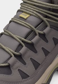 Κοντινή εικόνα γκρι παπουτσιού πεζοπορίας που παρουσιάζει υφή υφάσματος, επενδεδυμένη γλώσσα με μικρή κίτρινη ετικέτα και σφιχτά πλεγμένα δίχρωμα κορδόνια περασμένα μέσα από θηλιές.