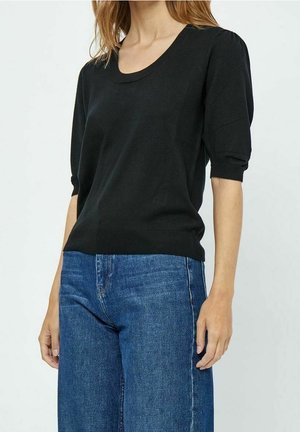 Femme portant un pull noir à manches courtes et un jean bleu taille haute, debout devant un fond clair uni.