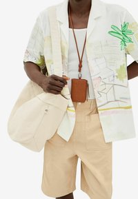 Camicia a maniche corte con design stampato colorato, pantaloni corti in cotone beige, borsa in tela color sabbia e porta carte in pelle marrone appeso a un cordino.
