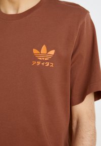 Brun bomull T-shirt med en orange Adidas-logotyp och japansk text. Korta ärmar, rund hals, slät yta, avslappnad passform.