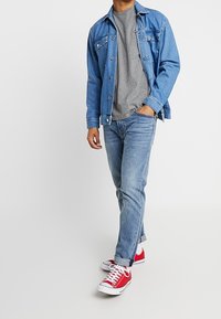 Chaqueta de mezclilla azul sobre una camiseta gris, combinada con vaqueros azul claro y zapatillas rojas. Ajuste casual con detalles de botones y puños remangados.
