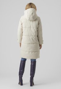 Manteau long matelassé beige avec une capuche, comportant des sections matelassées horizontales, porté sur un jean bleu et des bottes violettes jusqu'au genou.