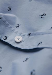Camisa azul clara con pequeños patrones de cangrejos negros. Presenta un botón blanco en el cuello y una tela suave con una ligera textura.