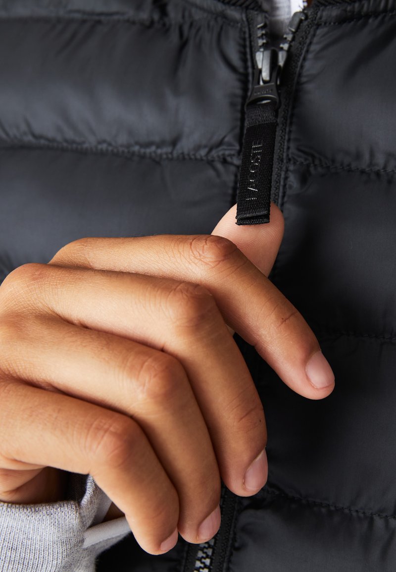 Doudoune noire avec texture matelassée, dotée d'une fermeture zippée et d'une tirette de zip à logo, avec une main nue saisissant le zip.