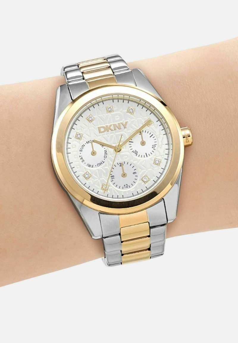 Zilveren en gouden DKNY polshorloge met witte wijzerplaat, drie subwijzers voor dag, datum en 24-uurs tijd, gedragen aan de linkerpols.