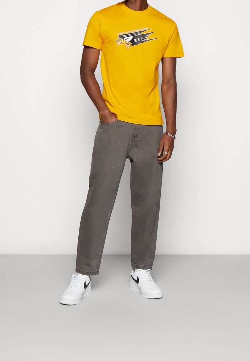 Żółta koszulka z krótkim rękawem z czarnym wzorem graficznym, zestawiona z szarymi spodniami o prostych nogawkach i białymi trampkami z czarnymi akcentami.