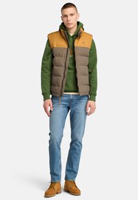 Padded vest in olive green and tan, featuring a zip closure, stand collar, and logo. Worn over a green hoodie, paired with blue jeans and tan boots.