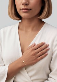 Goldene Armbanduhr mit einem Kreuzanhänger, ausgestattet mit einer strukturierten Kette und getragen an einer Hand über einem weißen Kleidungsstück. Glatte, neutralfarbene Nägel.