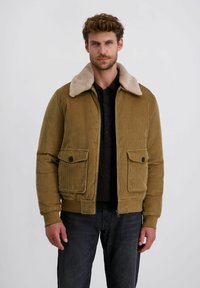 Olijfgroene corduroy jas met een beige nepbont kraag, twee voorzakken en ribgebreide manchetten en hem. Draagt over een donker shirt.