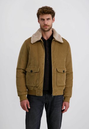 Olijfgroene corduroy jas met een beige nepbont kraag, twee voorzakken en ribgebreide manchetten en hem. Draagt over een donker shirt.
