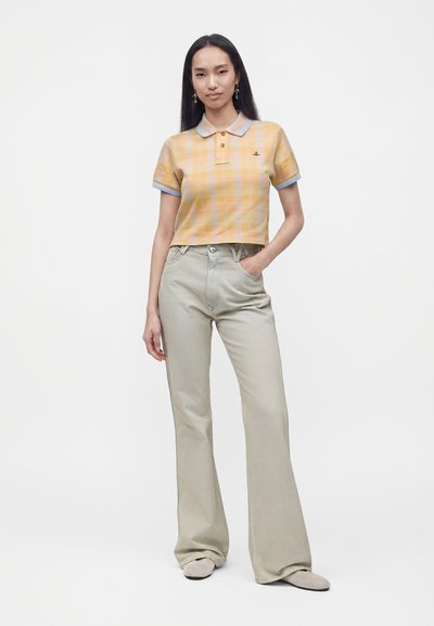 Femme aux longs cheveux foncés portant un polo jaune à carreaux à manches courtes et un pantalon large taille haute gris clair, debout avec une main dans la poche.
