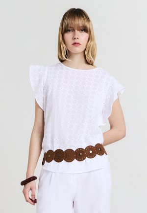 Giovane donna con capelli biondi lisci indossa una maglietta bianca a pizzo con maniche a volant, una larga cintura rotonda marrone, pantaloni bianchi e un braccialetto scuro.