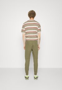 Pruhované bavlněné tričko v béžové, hnědé a černé barvě s krátkými rukávy, doplněné olivově zelenými tepláky a teniskami s bílými a zelenými detaily.