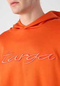 Felpa arancione con cappuccio, dal texture morbido, caratterizzata da un testo ricamato in rosa "targa" sul petto con un font corsivo.