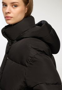 Czarna kurtka puchowa z wysokim kołnierzem i kapturem. Ma pikowane sekcje oraz gładką teksturę. Detale zapięcia są minimalistyczne i uproszczone.