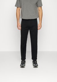 Pantaloni neri a taglio dritto, realizzati in materiale misto cotone. Abbinati a una camicia grigia a maniche corte e sneaker nere con motivo.