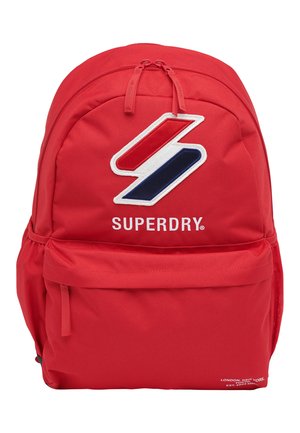 Sac à dos rouge avec fermeture zippée sur le dessus, arborant un grand logo blanc, bleu et rouge ainsi qu'une poche frontale supplémentaire. Fabriqué en tissu durable.
