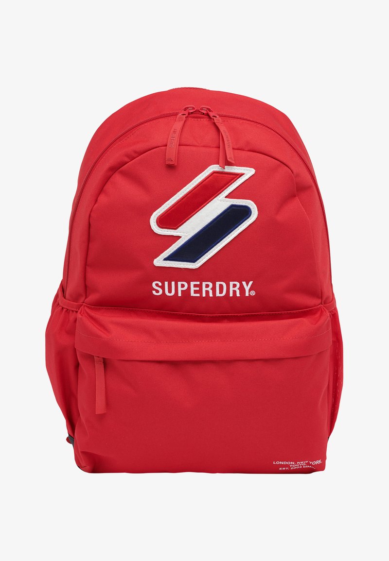 Sac à dos rouge avec fermeture zippée sur le dessus, arborant un grand logo blanc, bleu et rouge ainsi qu'une poche frontale supplémentaire. Fabriqué en tissu durable.