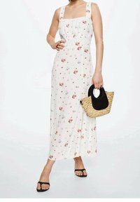 Robe maxi blanche à fleurs avec des bretelles épaisses, ornée de petits motifs de fleurs rouges et bleues. Tient un sac à main en osier avec des accents noirs.