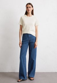 Beige T-Shirt mit kurzen Ärmeln, kombiniert mit blauen weit geschnittenen Jeans und braunen Sandalen, vor einem neutralen Hintergrund stehend.