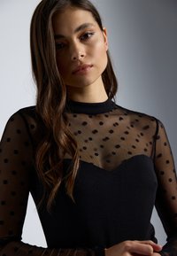 Haut noir ajusté avec une superposition en mesh transparent à pois, col montant et manches longues ; tissu lisse en dessous avec une découpe en forme de cœur.