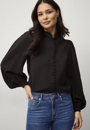Kvinde med langt mørkt hår iført en sort button-up bluse med flæser og puffede ærmer kombineret med blå denimjeans.
