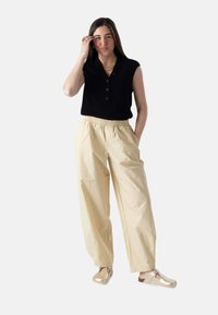 Mujer de pie con una mano tocándose el cabello, vistiendo una camiseta sin mangas negra, pantalones beige de pierna ancha y zapatos beige sin cordones.