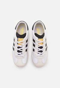 Vita sportskor med svarta ränder, texturerad svart tunga, gul logotyp och vita snören. Mjukt övermaterial och böjd sula.