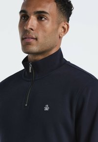 Marineblauwe ritsvest met hoge kraag, gemaakt van een zachte stof, met een klein wit pinguïnlogo op de borst.