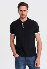 Camisa polo negra con cuello redondeado, que presenta tres botones blancos y detalles blancos en los dobladillos de las mangas. Textura de tela suave y lisa.