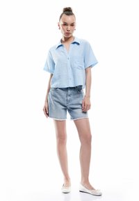Lysblå kortærmet skjorte med knapper og lomme, parret med lyse denimshorts med frayed kanter og hvide flade sko.