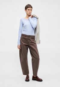 Svetlomodrá košeľa s dlhými rukávmi, voľné hnedé nohavice s vreckami a tmavohnedé topánky. Modelka drží svetlý pruhovaný blejzer na pleci.