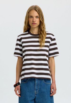 Femme aux longs cheveux blonds ondulés portant un t-shirt à manches courtes rayé marron et blanc et un jean bleu, debout devant un fond uni.