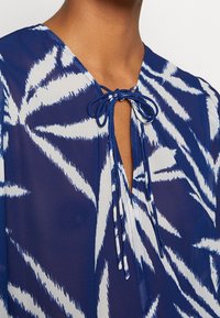 Blouse à motif bleu et blanc avec un col en V noué avec de fines lanières de tissu, laissant apparaître le haut de la poitrine et la zone du cou.