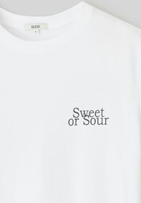 T-shirt en coton blanc avec un col rond et des manches courtes. Présente l'inscription « Sweet or Sour » imprimée en police stylisée foncée sur le devant.