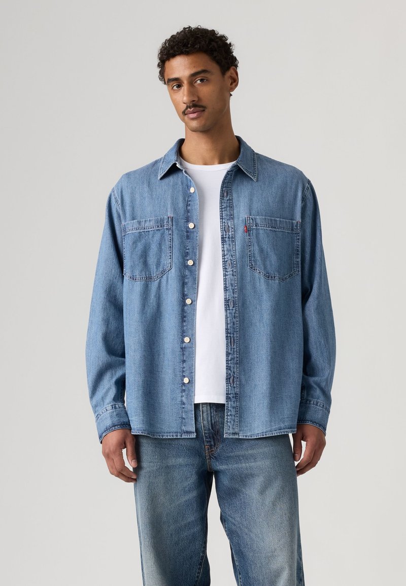 Homme portant une chemise en jean bleu clair boutonnée sur un t-shirt blanc et un jean bleu, debout devant un fond clair uni.