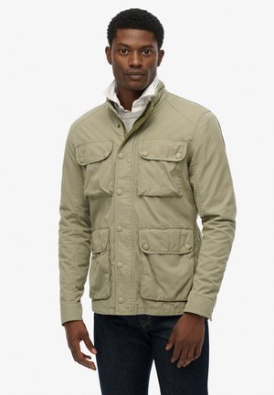 Helle olivgrüne Jacke mit vier Fronttaschen, Druckknöpfen, Stehkragen und einer glatten, matten Textur. Über einem grauen Pullover getragen.