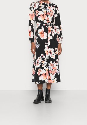 Zwarte bloemenjurk met roze en witte bloemen, knoopsluiting aan de voorkant, lange mouwen, ceintuur in de taille, gedragen met zwarte enkellaarzen.