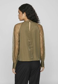 Blouse vert olive avec des manches en dentelle transparente, détail en trou de serrure au dos et manches longues, présentant un tissu doux et une coupe structurée.