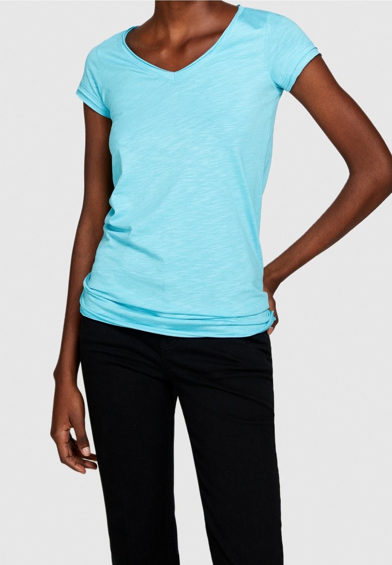 T-shirt bleu clair à manches courtes et en V, fabriqué en tissu doux et extensible, offrant une coupe décontractée et une légère texture. Porté avec un pantalon noir.