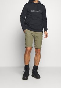 Felpa nera con cappuccio e tasca frontale, pantaloncini cargo verde oliva e scarponi da trekking neri. Abbigliamento casual da esterno.