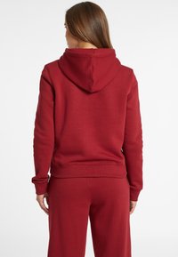 Vrouw met lang bruin haar, gekleed in een eenvoudige rode hoodie en bijpassende rode broek, met haar rug naar de kijker tegen een witte achtergrond.