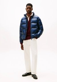 Granatowa pikowana kurtka puchowa o błyszczącym wykończeniu, z poziomymi przeszyciami, przednim zamkiem błyskawicznym i ściągaczami na mankietach, noszona na bordowym swetrze.
