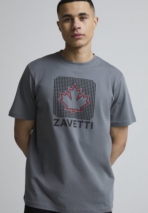 Junger Mann in grauem T-Shirt mit schwarzem Punktquadrat, rot umrandetem Ahornblatt und dem Schriftzug "ZAVETTI" auf einem einfarbigen Hintergrund.