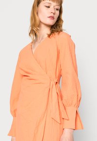 Vestido envolvente pêssego em coral suave com mangas longas, um decote em V profundo e faixa na cintura. Tecido texturizado com detalhes de pregas subtis.