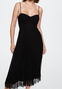 Černé krajkové šaty s tenkými ramínky, výstřihem do tvaru srdce a plisovanou sukní. Látka má květinový vzor a lemování, které sahá do poloviny lýtek.