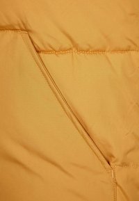 Gul pufferjacka i närbild, med slät tyg, diagonal söm och en dold ficka. Texturerad och lätt med en matt yta.