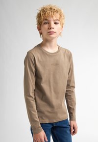 T-shirt a maniche lunghe in cotone marrone chiaro con scollatura rotonda e dettaglio del logo sul petto, abbinato a jeans blu in denim.