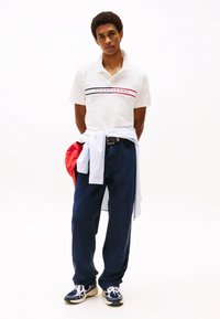 Camisa polo blanca con rayas azules y rojas, pantalones caqui y zapatos navy. Los accesorios incluyen una chaqueta roja y un suéter blanco atado a la cintura.