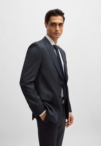 Dark gray suit with a textured fabric, single-breasted design, notched lapels, and matching tie. Model strikes a side profile pose.